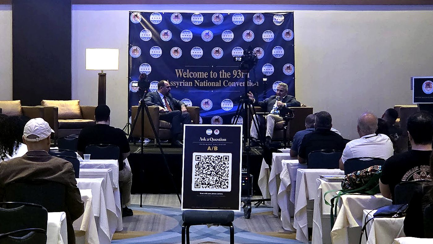 An Ask a Question sign with a QR code standing in the aisle of a convention room, panel on stage behind it